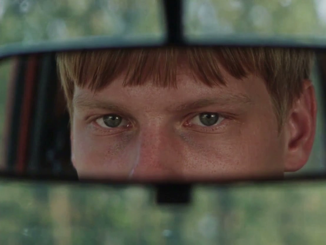 One continuous shot, no cuts. Extreme close-up of a young man's brow, eyes and bridge of the nose seen as a reflection in a narrow horizontal strip of glass, a car rearview mirror, framed above and below by dark metallic borders. He has short blond hair with straight bangs falling over the forehead, fair skin and light-coloured eyes, matching the person in the reference. Eyes centered in the strip at eye level, sharp focus on the eyes and brow, warm catchlights in the irises. Outside the glass, soft out-of-focus green foliage and gentle daylight.

Timing: 0.0 to 1.0 s he stares intently and steadily into the mirror, vigilant and tense. At about 1.0 to 1.25 s a subtle gaze shift: the eyelids narrow slightly, the gaze drops downward and slightly away, with a barely perceptible tilt of the head downward. From about 1.25 s to the end of the clip he holds this lowered, off-axis line of sight, still and tense, conveying hesitation and inner tension.

Camera: locked-off camera on the mirror. The subject moves inside the frame. No pan, tilt, push, pull, orbit, drift, zoom, or reframing. End view is the same framing with the gaze lowered, held.

Look: soft naturalistic daylight with low-key car-interior shadows, warm skin tones, rich contrast, heavy 35mm film grain, vintage 1990s analog thriller look. Silent, no dialogue, no music. Show only the reflection in the mirror strip; do not reproduce the reference's layout, panels, white background, or full-body views, and add no text.