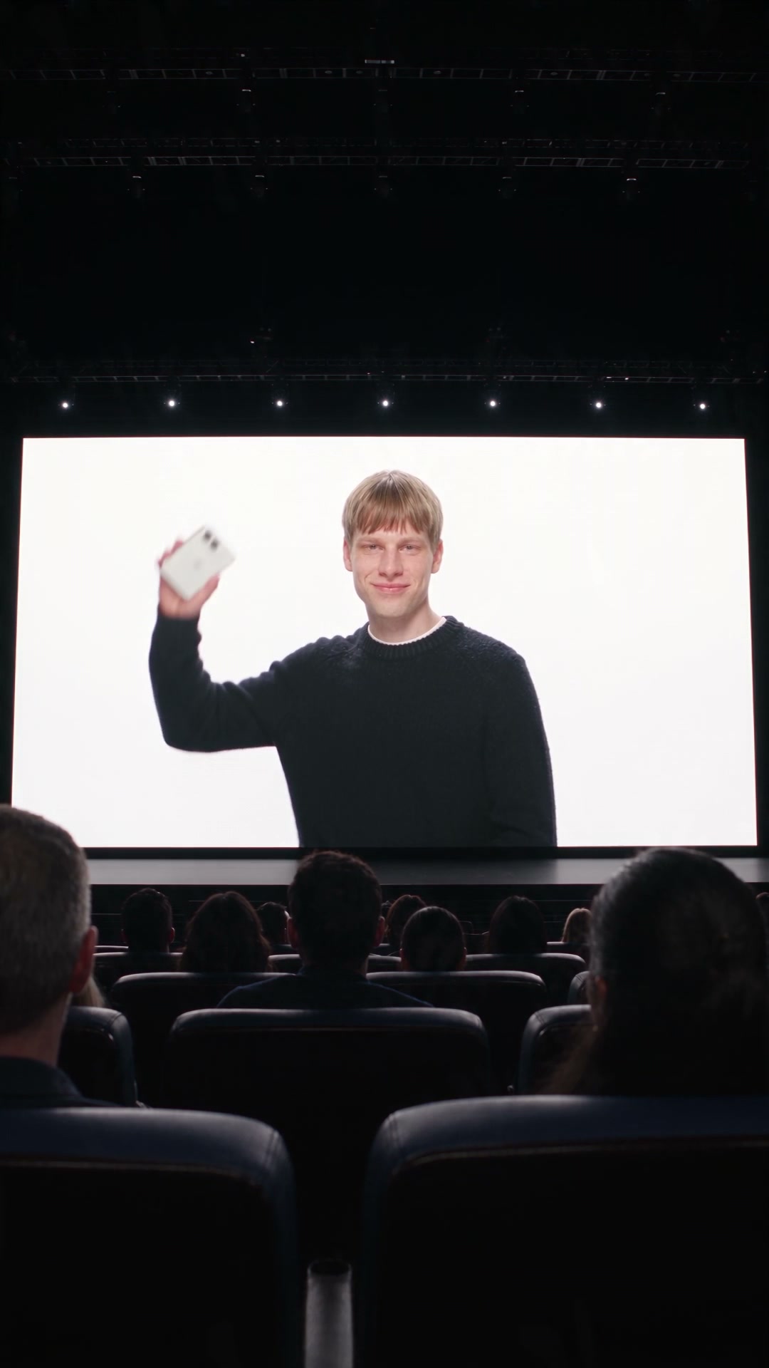 Camera Angle & Perspective:

Strict first-person perspective (POV shot). A single, continuous fluid camera movement with zero cuts, simulating a human eye view. Hand motion must maintain realistic human anatomy: exactly five fingers, correct joint proportions, natural wrist rotation, and no floating or warping artifacts.

Setting & Environment:

Inside a dimly lit, high-end tech conference auditorium filled with a seated audience. In the background, a massive glowing presentation screen displays a male speaker in a black sweater against a clean white backdrop.

Action Breakdown (Second-by-Second Flow):

The Toss (0:00 - 0:02):

From the POV angle, look toward the large illuminated stage screen. On screen, the male presenter smiles, raises a sleek light-gray folded smartphone, and throws it forward toward the camera in a smooth motion.

Mid-Air Flight & Unfolding (0:02 - 0:04):

The camera tilts slightly up as the physical foldable phone flies out from the screen area, spinning through the dark air of the auditorium. Mid-flight, the device smoothly unfolds 180 degrees into a wide inner screen displaying a clear iOS-style home interface with vibrant app icons, a photo widget, and a clock.

The Catch & Audience Shock (0:04 - 0:07):

The camera moves downward to track the landing. A single, perfectly rendered right human hand reaches up into the lower frame. The open smartphone drops seamlessly and gently into the palm. Adjacent audience members in the surrounding seats turn their heads toward the hand in genuine shock and jaw-dropping amazement, laughing and reacting naturally.

Inspection & Folding (0:07 - 0:10):

The hand holds the unfolded phone steady for a fraction of a second, showing the sharp screen display. The hand then closes the phone shut with a firm, clean snap. The camera focuses on the folded exterior: a matte off-white rear glass casing with a dual-camera module on top and an Apple logo centered.

Lighting, Texture & Rendering Style:

Photorealistic 8K render, dramatic cinematic key lighting from the main stage, natural motion blur during the aerial throw, shallow depth of field focusing sharply on the handheld phone while keeping the background audience slightly soft. Clean geometric symmetry on the smartphone body, zero texture flickering or digital distortion. High frame rate (60fp Source B