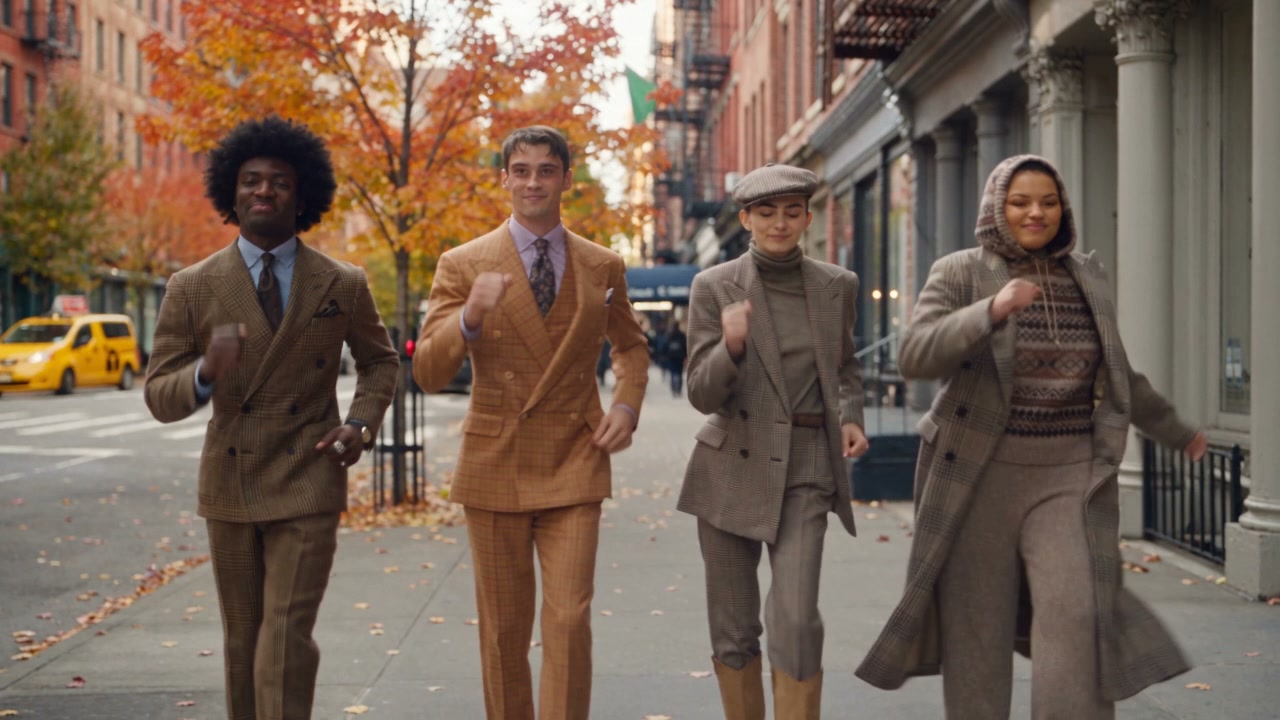 Four friends, Dancer 1, Dancer 2, Dancer 3 and Dancer 4, dance together on a New York City sidewalk on a fall day. Each dancer's face, hair, skin tone and body must match their own identity reference exactly, with no blending between people: the first four reference images are the only source of faces and hair, and the last four images show clothing only (their faces are not used).

Dancer 1: man with a full rounded black afro, small stud earring, large silver ring. Dancer 2: lean athletic man with short dark-brown hair and a side-swept fringe, light grey-green eyes, angular jaw, tanned skin; his hair is completely dry, soft and matte, with no wet look, no water, no sheen, and he wears no necklace. Dancer 3: woman with light-brown hair slicked straight back with a side part (not curly), strong dark brows, grey-green eyes, laughing as she dances. Dancer 4: woman with dark-brown hair in a loose messy updo, warm tan skin, curvy build, layered gold necklaces with a nameplate pendant.

Wardrobe (clothing only from the last four images): Dancer 1 in the brown glen-plaid double-breasted suit, light blue shirt and patterned tie. Dancer 2 in the olive/tan windowpane double-breasted suit, lavender shirt and tie. Dancer 3 in the newsboy cap, grey houndstooth double-breasted blazer, grey turtleneck, houndstooth trousers and tan knee boots. Dancer 4 in the hooded fair-isle knit, grey-brown houndstooth long coat and grey wool trousers. No handbags, folios or held props; hands free for dancing.

Setting: a wide Manhattan sidewalk lined with brownstones and storefronts on a bright autumn day, yellow cabs softly blurred in the distance, orange, red and golden trees, fallen leaves on the pavement, crisp soft daylight. Only a few blurred pedestrians far in the background. Exactly four dancers in the foreground.

Shots, with hard cuts between them and camera movement only inside a shot:
Shot 1 (0-4s): locked-off medium-wide knee-up shot at eye level, the four dancers in a loose row facing camera, all four faces clearly visible, dancing joyfully with bouncy steps and arm waves. No camera movement.
Cut to Shot 2 (4-7s): medium shot from a slightly low angle, faces clearly visible and sharp, smooth sideways track along the row as each dancer does a quick signature move (spin, hair flip or shoulder shimmy) and the coats swing.
Cut to Shot 3 (7-10s): medium-wide shot, slow push-in while all four bounce together, then strike a happy group pose and hold it for the last half second.

Style: loose, energetic fashion-dance video with hair tosses, arms up, shoulder shimmies, playful poses, laughter and buoyant group energy, similar to a casual studio dance montage but outdoors. Keep each dancer's identity and outfit consistent in every shot.

Audio: upbeat, catchy instrumental background music throughout, pop-funk groove with claps and bright guitar, plus light street ambience. No speech, dialogue, singing or lip syncing. No captions, subtitles, titles, labels or watermarks.