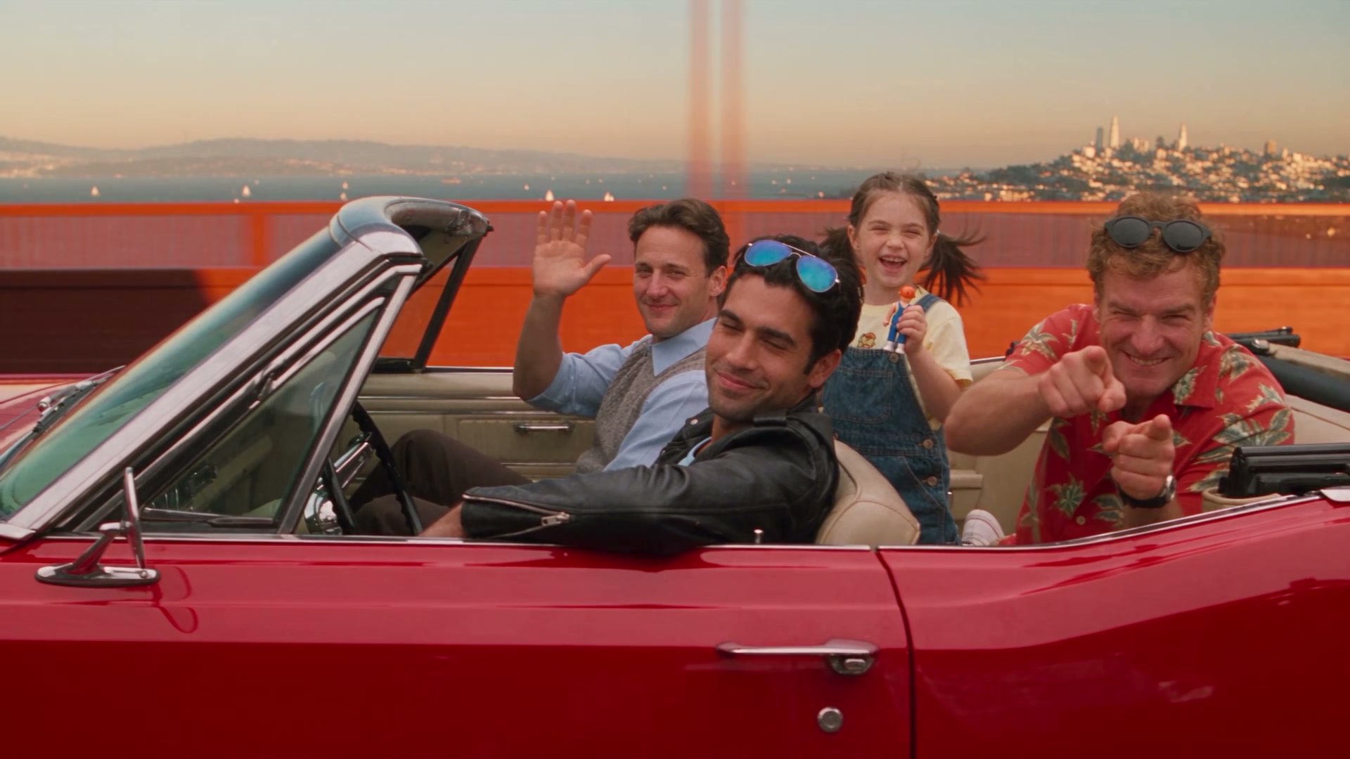 Car-to-car tracking shot running parallel to a cherry-red vintage convertible with a tan interior, top down, crossing the Golden Gate Bridge left to right, the bridge cables sweeping past behind it, sailboats on the bay far below, warm golden late-afternoon light. Standard normal lens character with a 47-degree diagonal field of view, held constant with no field-of-view drift. Dex drives, relaxed, one wrist resting on the wheel. Pete sits in the front passenger seat and waves warmly straight at the camera. In the back seat, Frankie kneels up on the seat waving with her small orange-haired toy figure in her fist, pigtails whipping in the wind, laughing; Marv points finger-guns at the camera. All four look directly at the camera and smile with open, direct opening-credits energy. Exactly these four people are in the car, no extras. Wind moves hair and collars naturally; the convertible rides with real suspension sway.