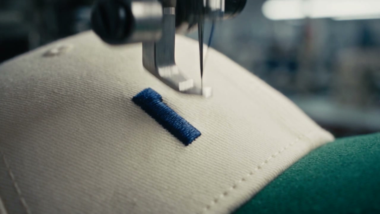 Cinematic 15-second commercial video sequence showing the precision manufacturing of a cream and green cap, shot on 35mm film with sharp focus, warm natural daylight, and dynamic camera transitions. Aspect ratio 16:9, 15 seconds duration.

[00:00 - 00:04] Extreme macro close-up of an automated industrial embroidery needle rapidly stitching dark blue thread into off-white canvas fabric, forming the text "MITO" alongside tiny embroidered tennis player figures near a dark green cotton brim.

[00:04 - 00:07] Close-up tracking shot of the heavy-duty machine finishing the "MITO" embroidery, pulling back to reveal the completed off-white and forest-green cap held on the machine mold inside a bright workshop.

[00:07 - 00:10] First-person perspective (POV) shot of a hand grabbing the cream and green "MITO" cap from the machine and tossing it forward into the air through an open doorway into the sunny outdoors.

[00:10 - 00:13] Seamless match-cut transition as the cap flips mid-air, gliding through the vibrant, aesthetic streets of Tenerife with palm trees, coastal sunlight, and pastel building facades in the background.

[00:13 - 00:15] The cap lands smoothly onto the head of a stylish, cool young woman with long dark hair, wearing an oversized plaid blazer, sitting outdoors in a sunny streetwear aesthetic as she adjusts the green brim with a confident, effortless smile. Final quick macro push-in on the embroidered "MITO" logo.